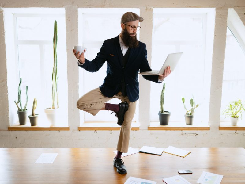 Meditation, calmness. Young bearded man, office clerk having fun, doing yoga on wooden table in modern office at work time with gadgets. Concept of business, healthy lifestyle, sport, hobby